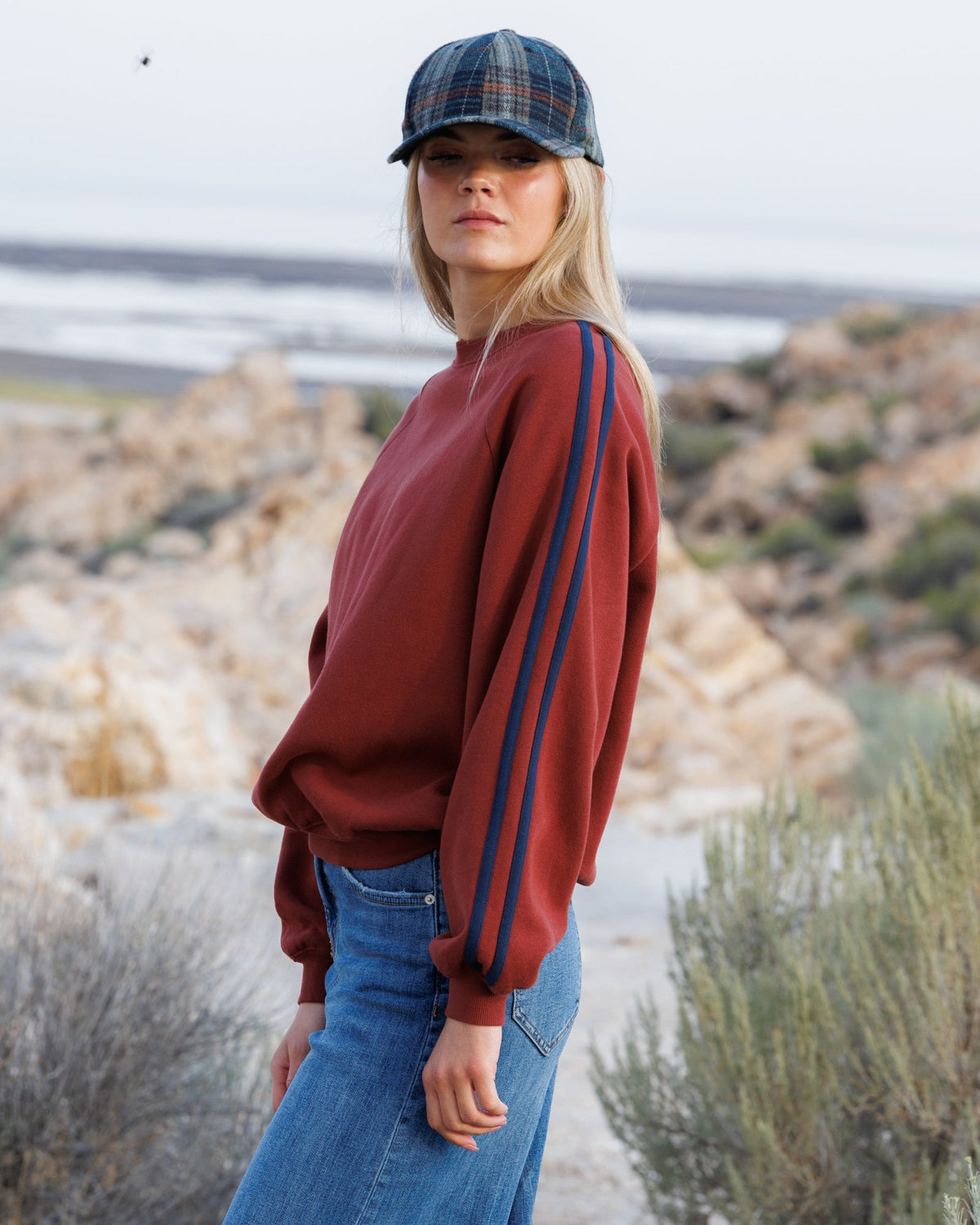 A woman wearing a rust-red crewneck sweatshirt with blue stripes down the sleeves, paired with blue jeans and a plaid flat cap, standing outdoors against a rocky, coastal background.