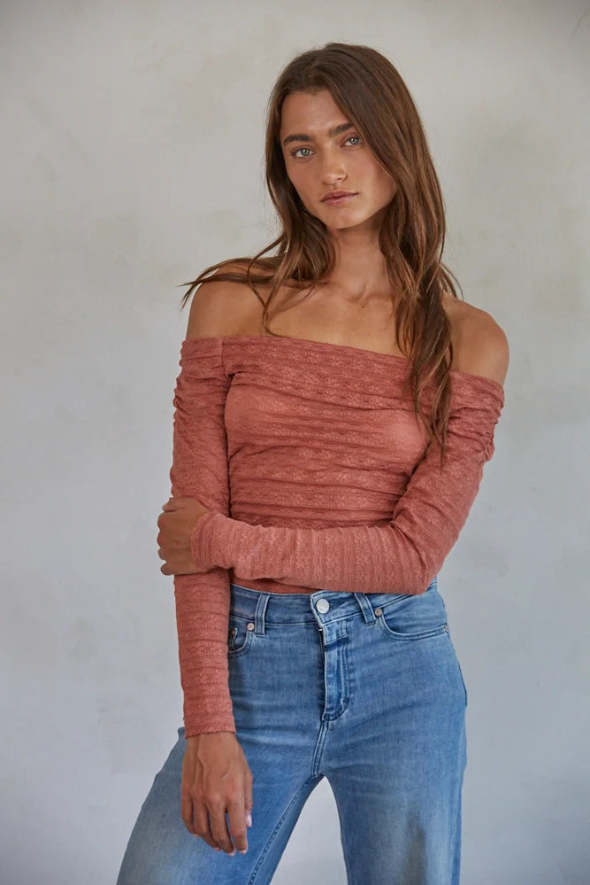 A woman wearing a pink one-shoulder long sleeve lace bodysuit with a flattering off-the-shoulder neckline, paired with blue jeans.