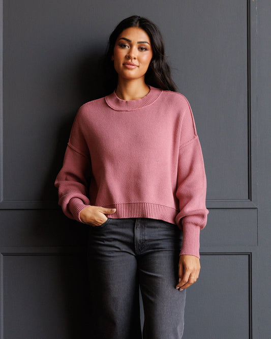 A woman wearing a dusty rose pink crewneck knit sweater and dark gray jeans, standing against a dark gray paneled wall.