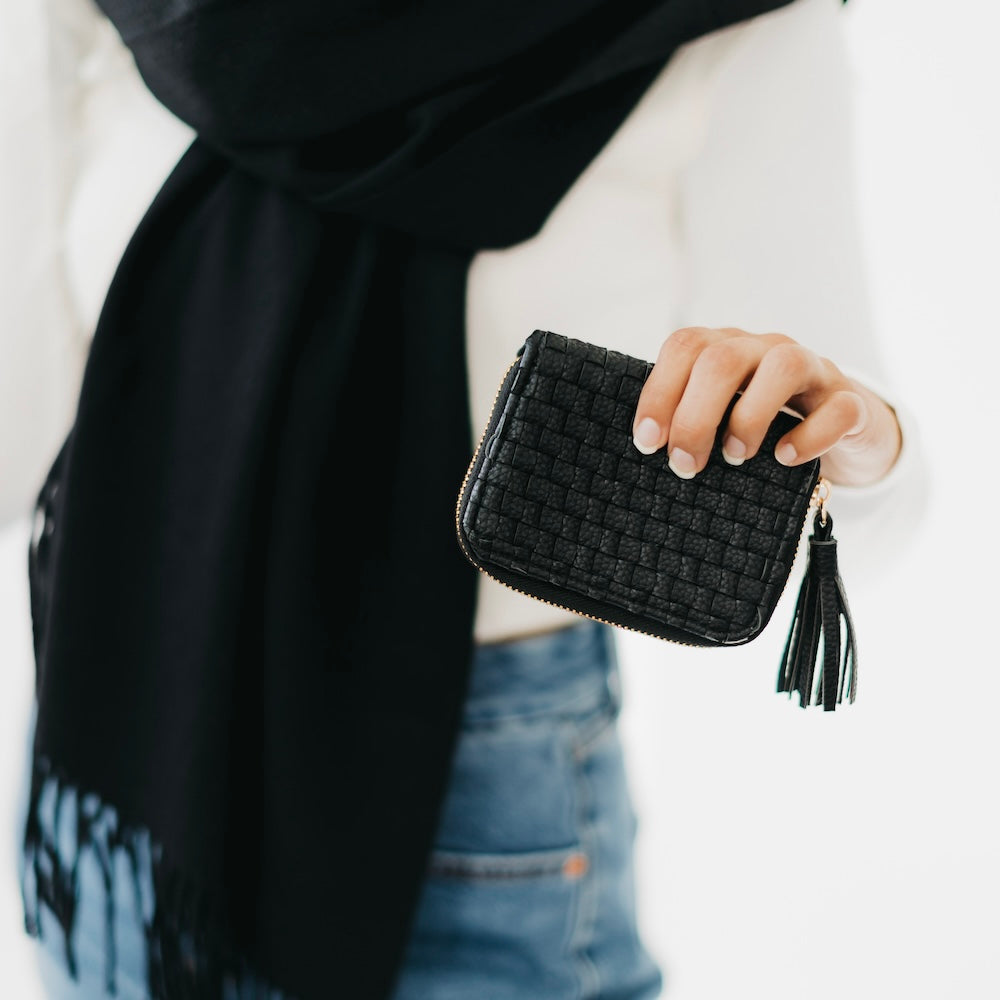 Person holding a black woven clutch with tassels against a neutral background