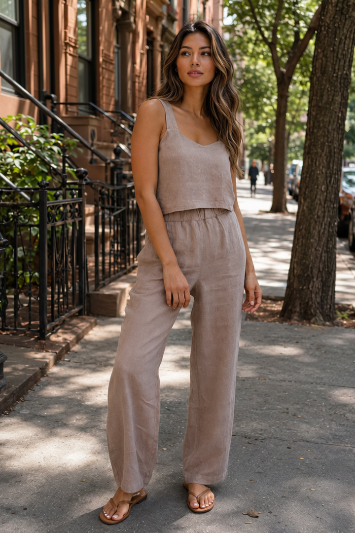 A woman stands on a city sidewalk wearing a matching sleeveless crop top and wide-leg pants set in a light taupe linen-like fabric, paired with tan sandals.