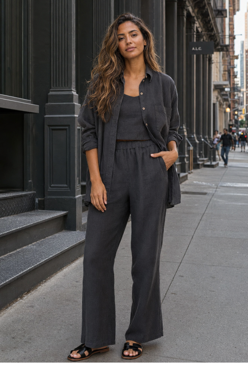 A woman stands on a city sidewalk wearing a dark charcoal gray matching lounge set consisting of a button-up shirt and wide-leg pants, styled with black slide sandals.