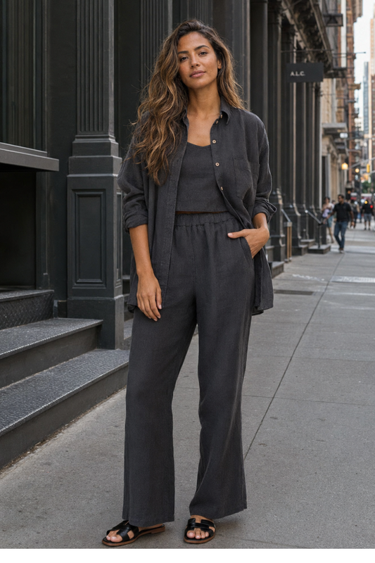 A woman stands on a city sidewalk wearing a dark charcoal gray matching lounge set consisting of a button-up shirt and wide-leg pants, styled with black slide sandals.