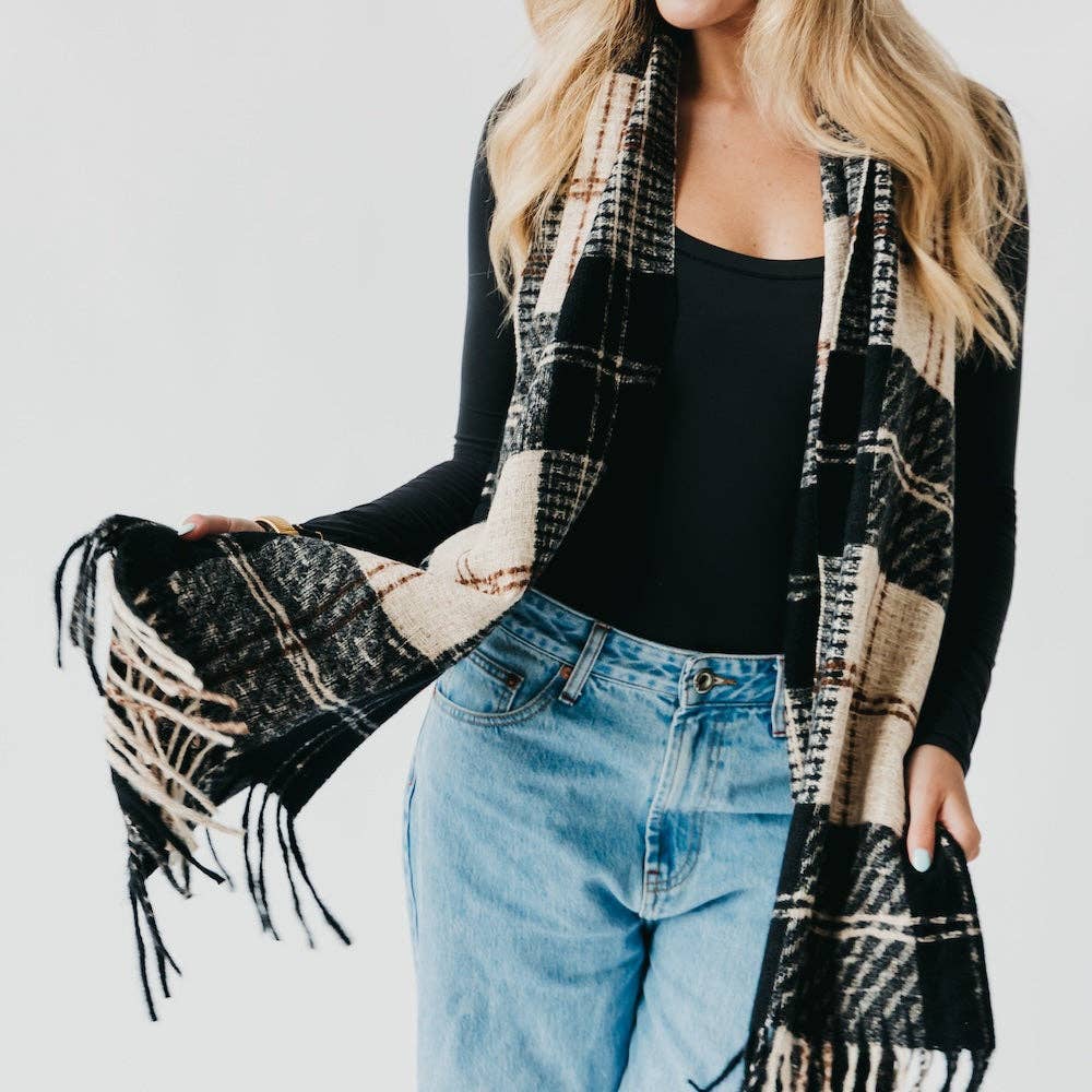 Person wearing a black and beige plaid scarf with tassels on a plain background