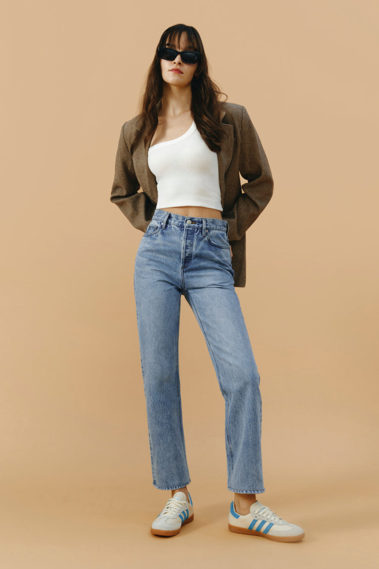 Woman wearing a white top, brown jacket, blue jeans, and sunglasses on a beige background