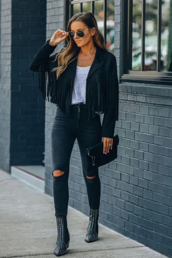 Woman wearing a black fringe vegan seude jacket, ripped jeans, and studded boots against a brick wall.