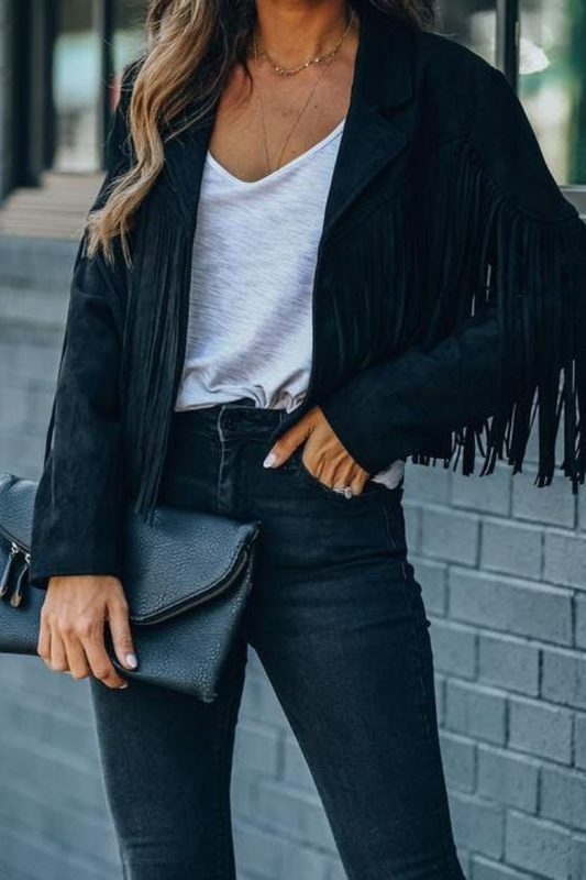 Person wearing a black fringe jacket, white top, and dark jeans, holding a black clutch against a gray brick wall.