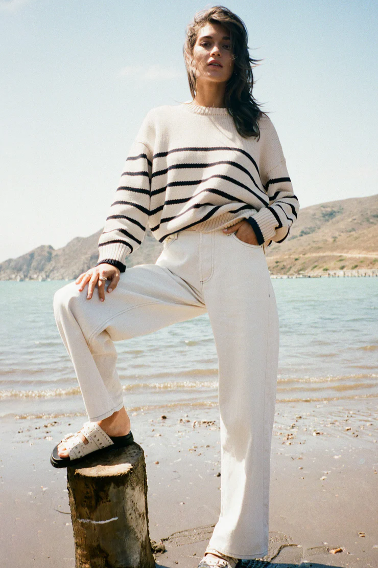 Woman in a striped sweater and white pants standing on a beach with mountains in the background