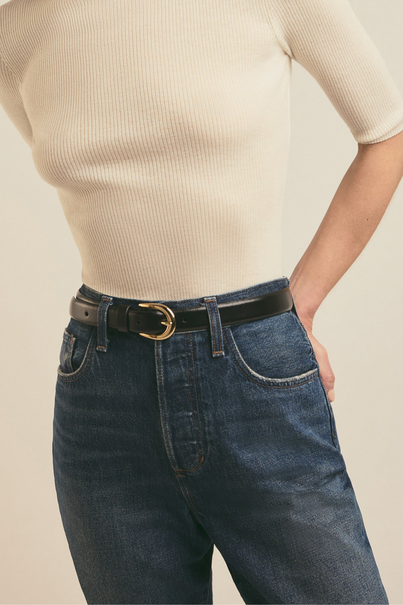 Person wearing a beige sweater and blue jeans with a black belt on a beige background