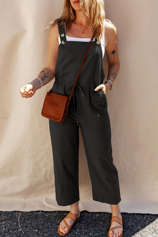 A woman wearing a sleeveless, buttoned strap overall in a solid color design, paired with a white tank top and carrying a brown bag.