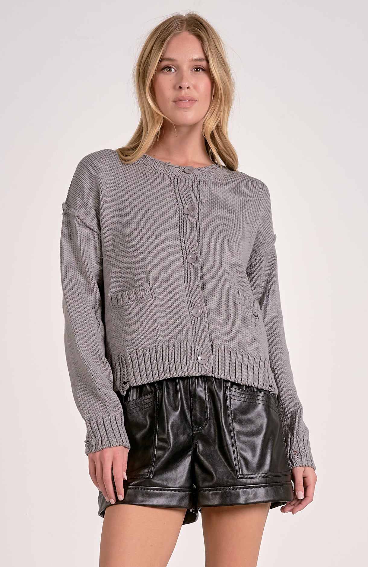 Woman wearing a gray knitted cardigan over a black top with leather shorts on a white background