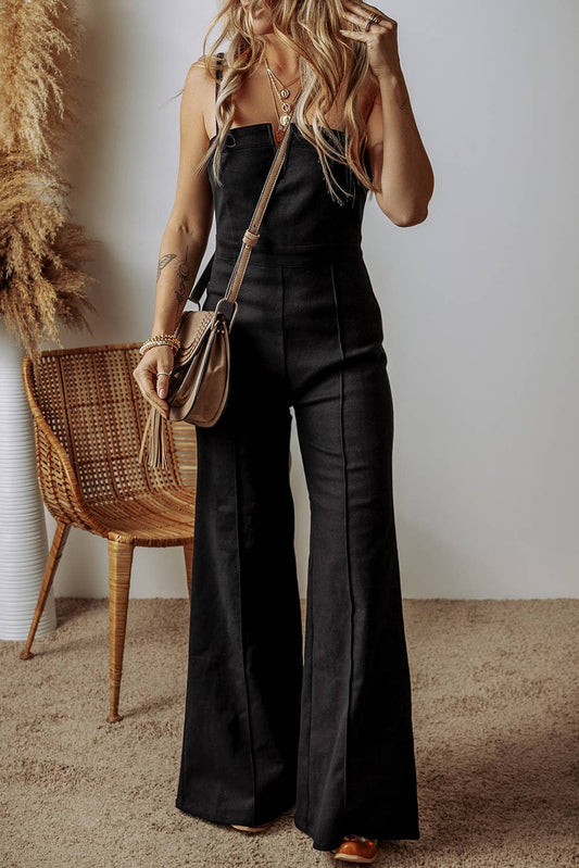 A woman wearing a black flare leg jumpsuit with a back zipper, paired with a straw bag and heels, standing indoors.