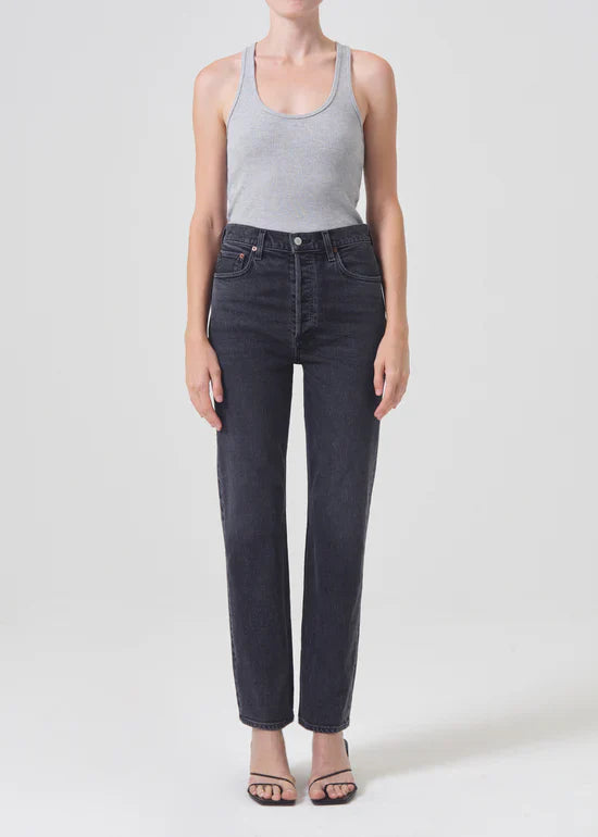 A model wearing Agolde High Rise Stovepipe jeans in the "Metal" wash. The jeans are a washed-down black, high-waisted, and have a slim straight leg that falls to the ankle.