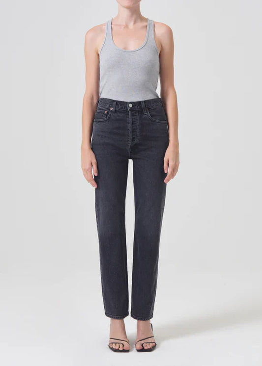 A model wearing Agolde High Rise Stovepipe jeans in the "Metal" wash. The jeans are a washed-down black, high-waisted, and have a slim straight leg that falls to the ankle.