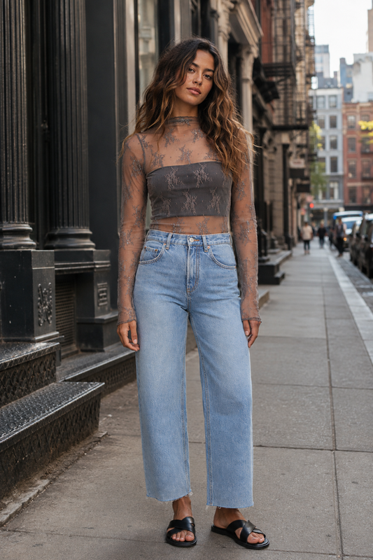 A woman stands on a city sidewalk wearing a sheer black lace crop top and light-wash wide-leg blue jeans, with black slide sandals. The background shows urban buildings and street details.
