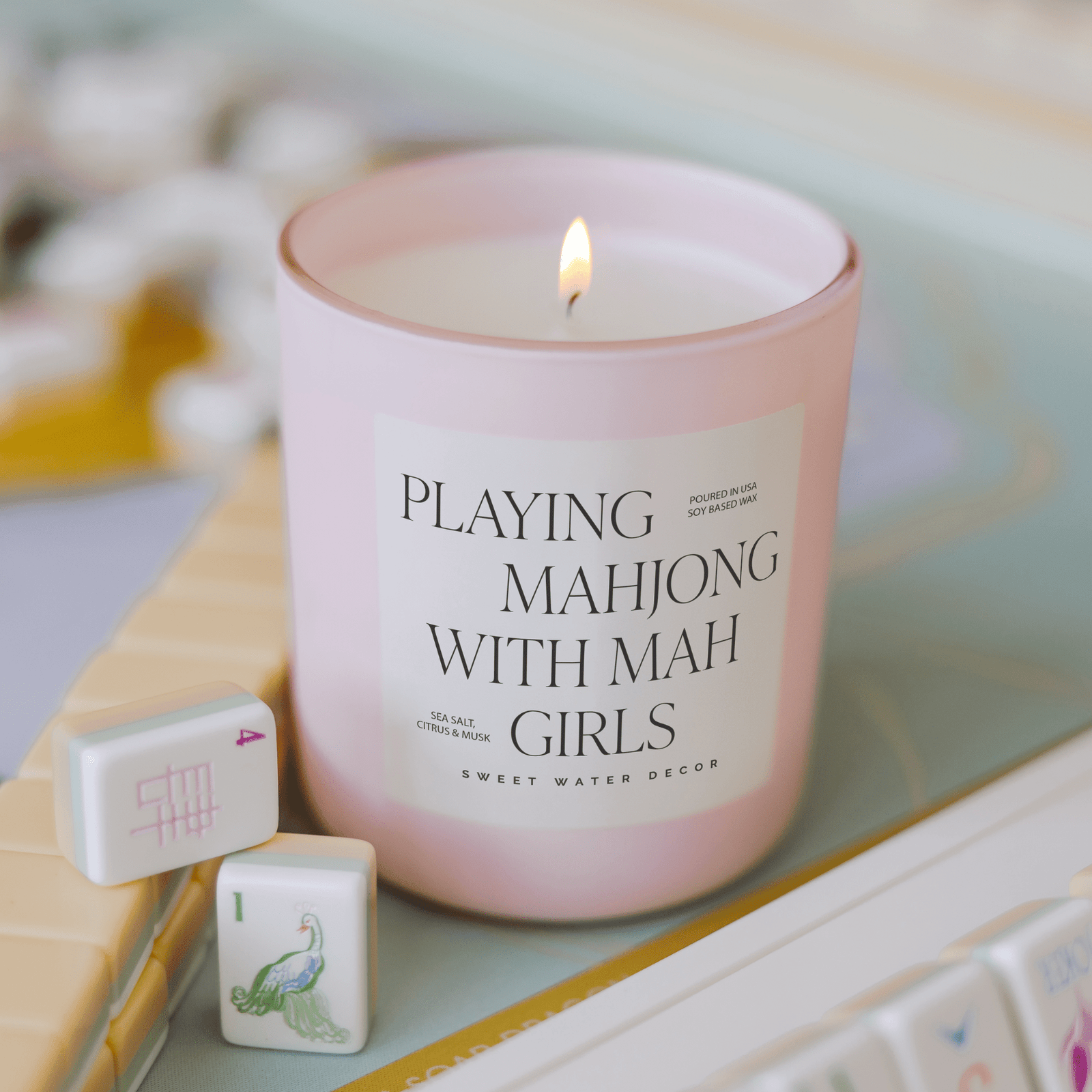 Pink candle labeled 'Playing Mahjong with Mah Girls' on a surface with mahjong tiles.