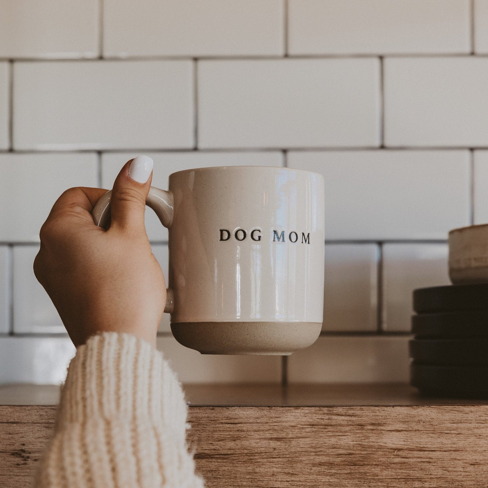 Hand holding a beige mug with 'DOG MOM' text in a kitchen setting.