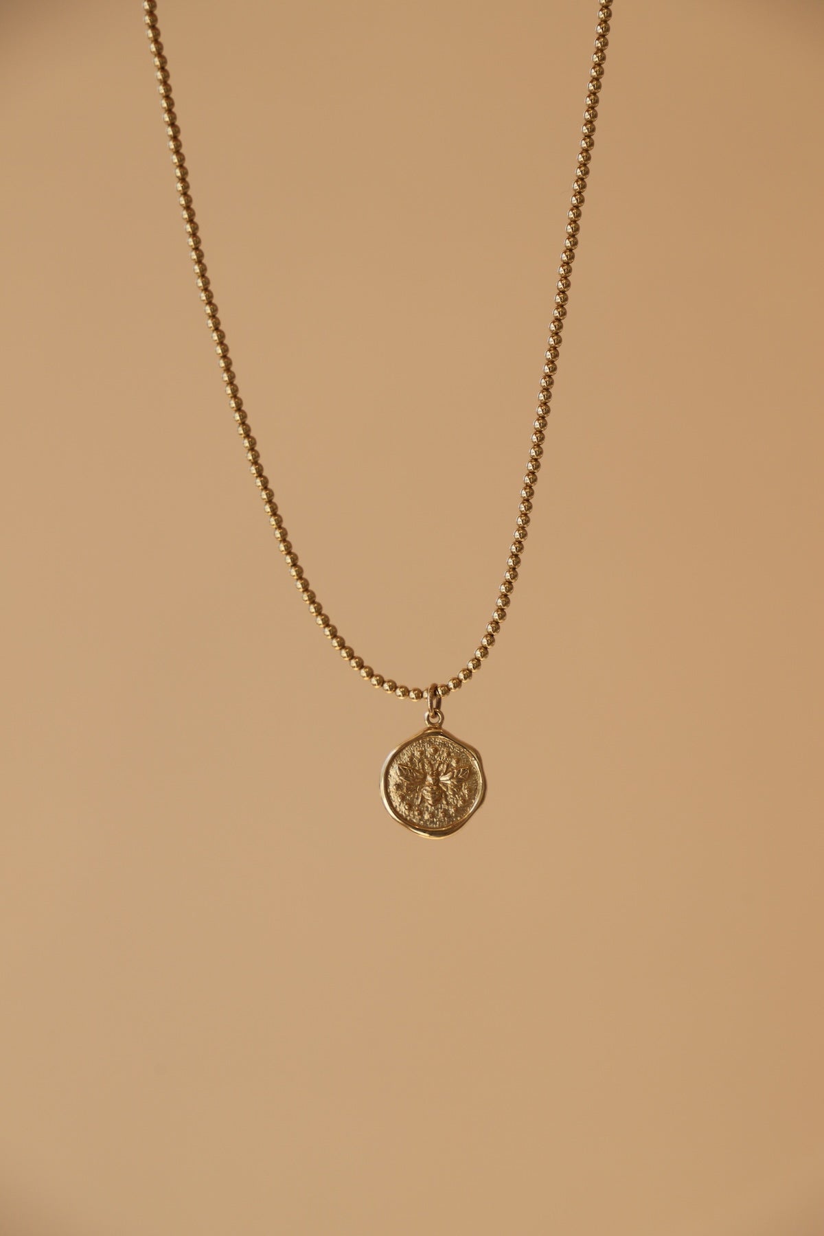 Gold necklace with a wax seal bee charm on a beige background