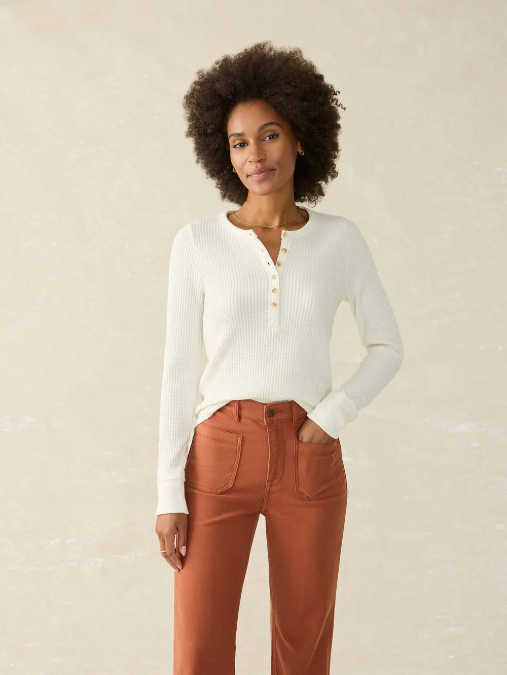 Woman wearing a white ribbed sweater and brown pants against a beige background