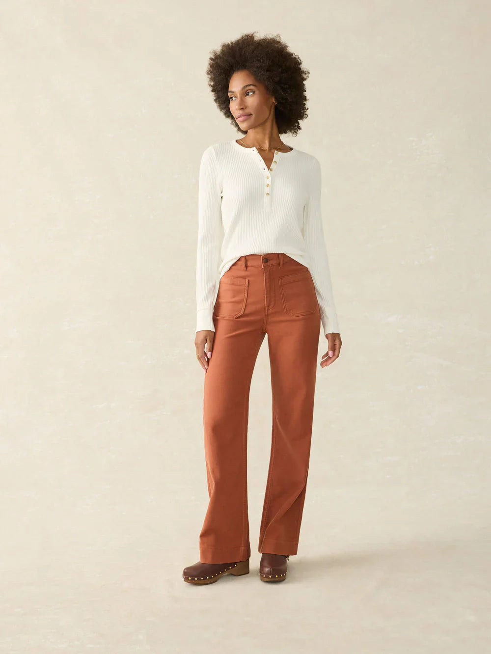 Woman wearing a white sweater and rust-colored pants on a beige background