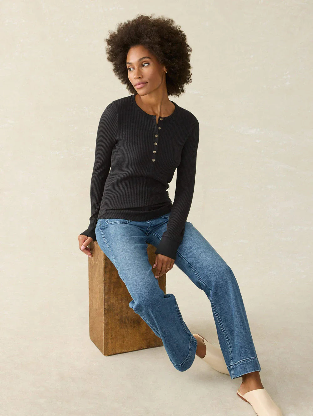 Woman wearing a black long-sleeve top and blue jeans sitting on a wooden block against a beige background