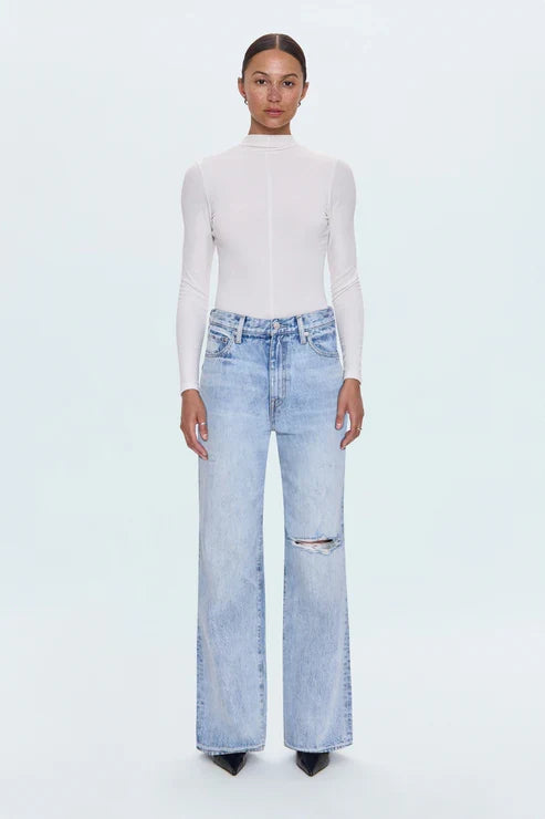 A model stands against a light gray studio background wearing a fitted white mock-neck long-sleeve top and light-wash blue wide-leg jeans with a distressed knee detail.