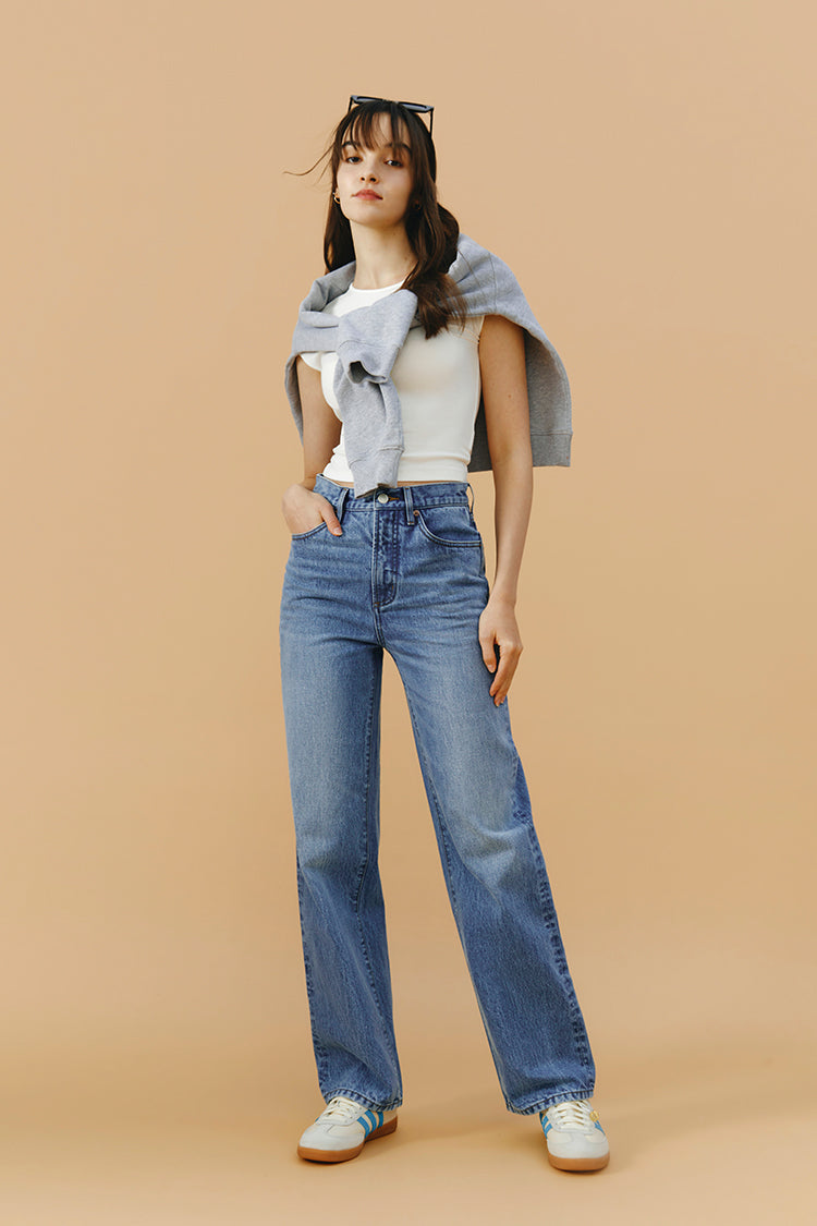 Woman wearing a gray sweater, white top, and blue jeans against a beige background