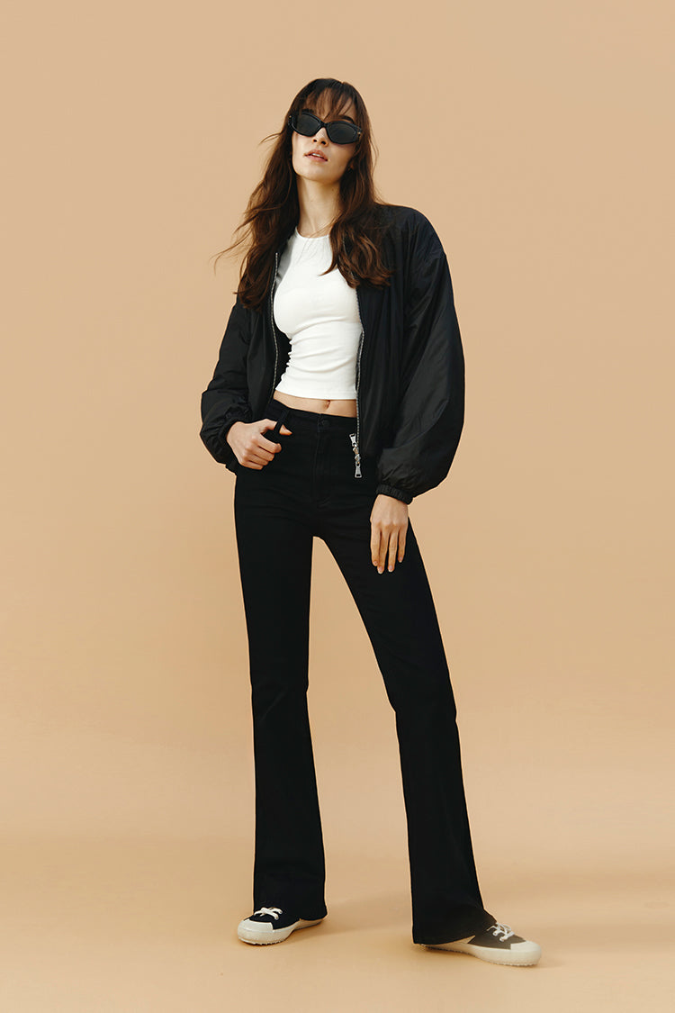 Woman wearing a black bomber jacket, white top, black pants, and sunglasses on a beige background