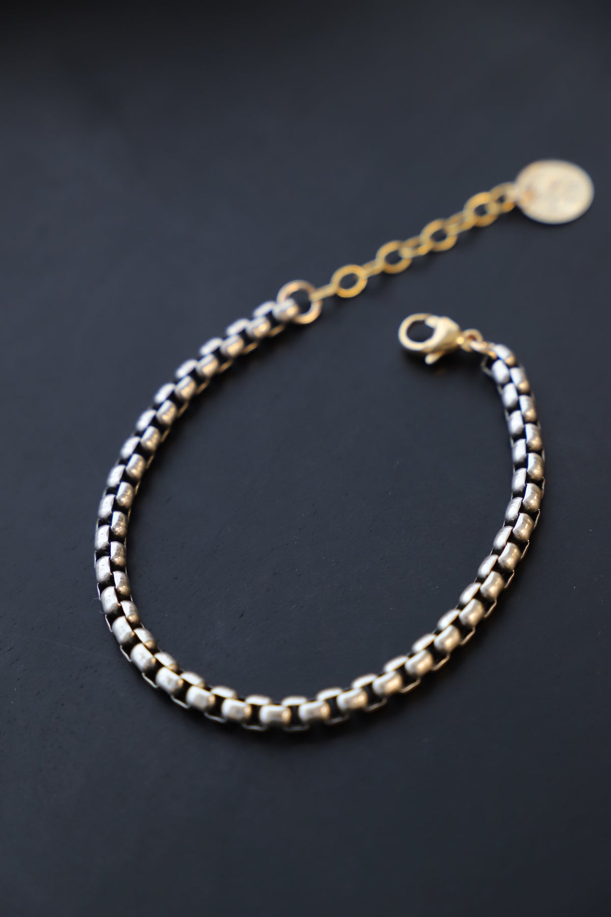 A close-up shot of the Nova Chain Bracelet, a two-tone accessory featuring a round silver box chain, a gold lobster clasp, and a small gold coin drop.