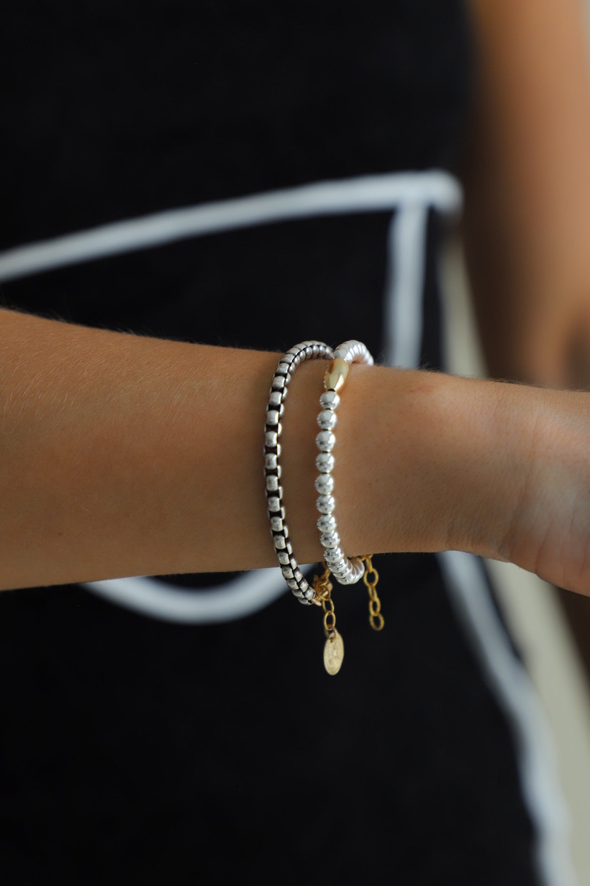 A person's wrist wearing the Nova Chain Bracelet, showing the contrast between the thick silver round box chain and the gold clasp and coin drop.

