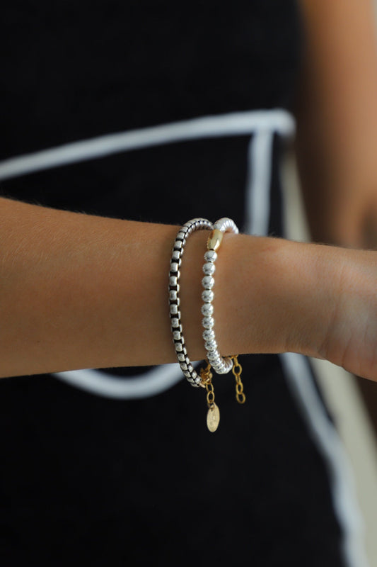 A person's wrist wearing the Nova Chain Bracelet, showing the contrast between the thick silver round box chain and the gold clasp and coin drop.

