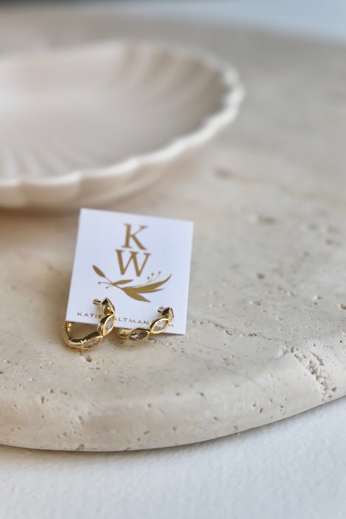 Gold earring on a white card with 'KW' branding on a beige surface