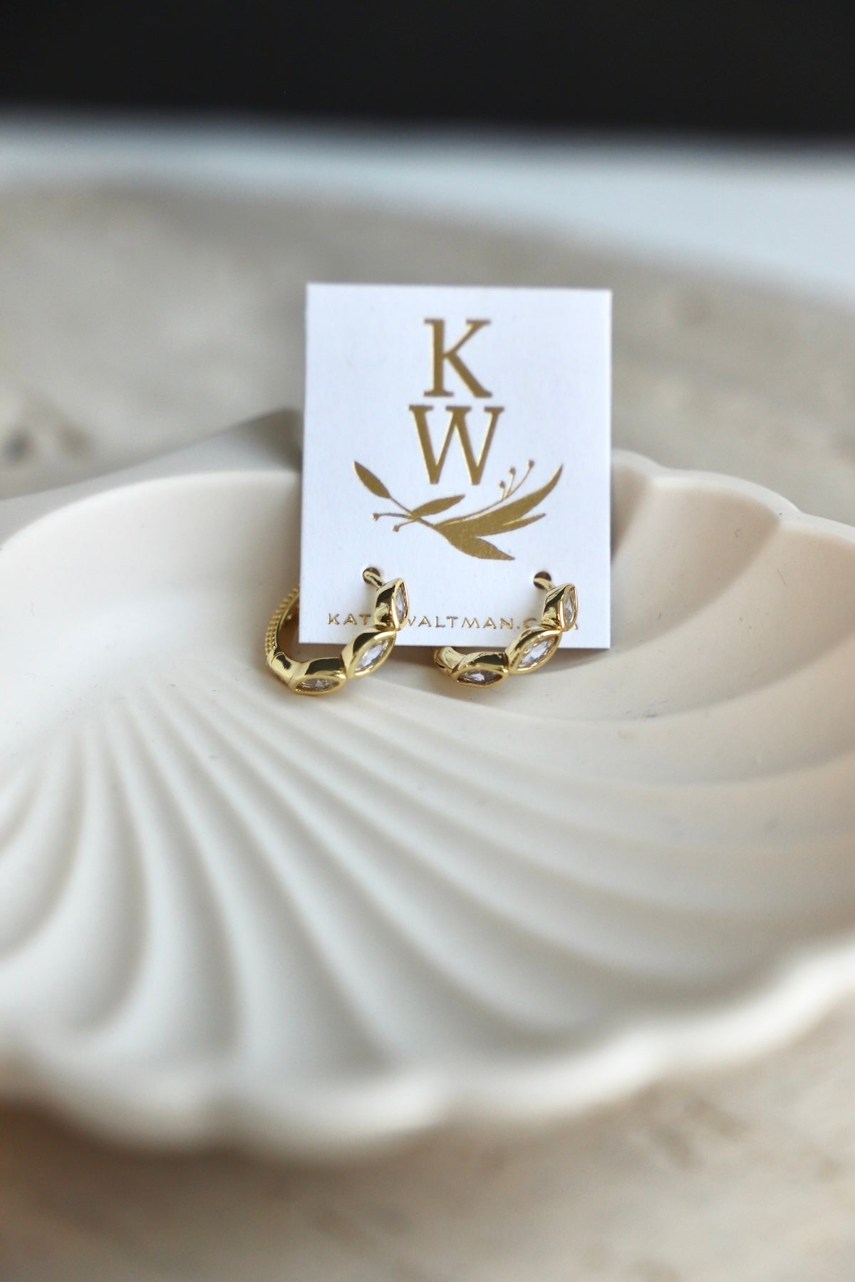 Gold earrings on a white card with 'KW' initials, placed on a shell.