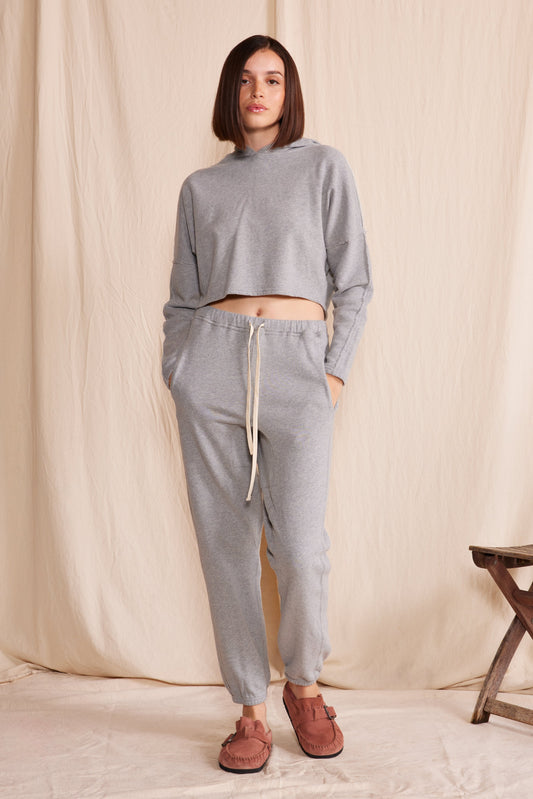 Woman wearing a heather gray cruz sweatpants and heather gray hooded sweatshirt with a wooden stool in the background