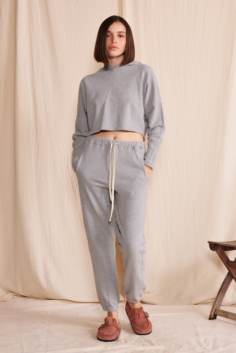 Woman wearing a heather gray cruz sweatpants and heather gray hooded sweatshirt with a wooden stool in the background