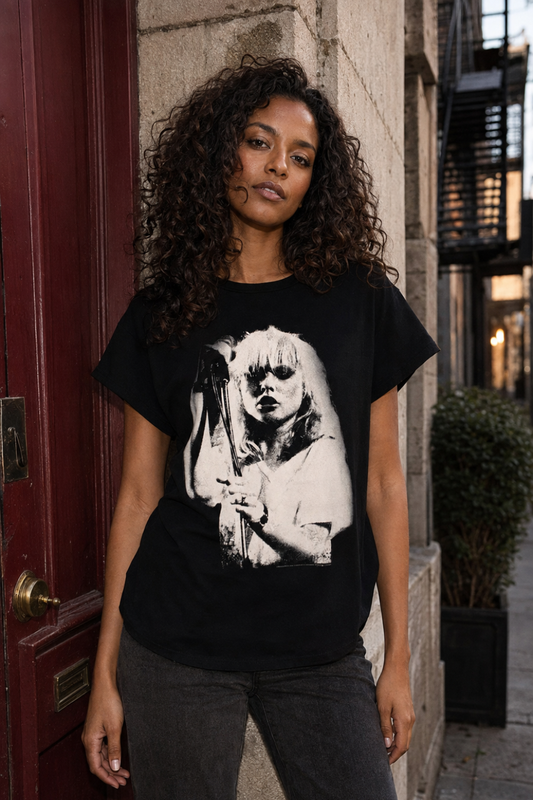 A woman with curly dark hair stands outdoors wearing a black graphic T-shirt with a large white high-contrast portrait print on the front, paired with dark gray jeans.