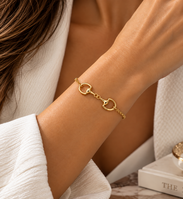 A close-up of a woman's wrist wearing a delicate gold horsebit chain bracelet, styled with a white textured robe.