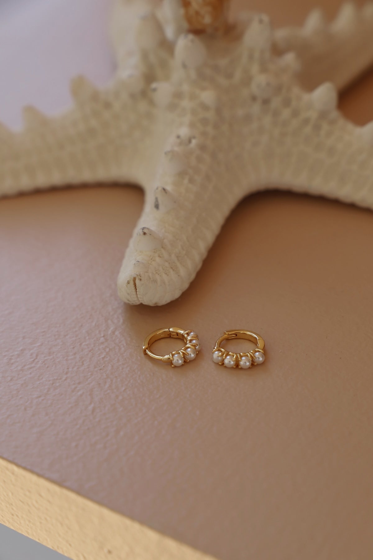 Gold hoop earrings with small pearls on a beige surface with a starfish.