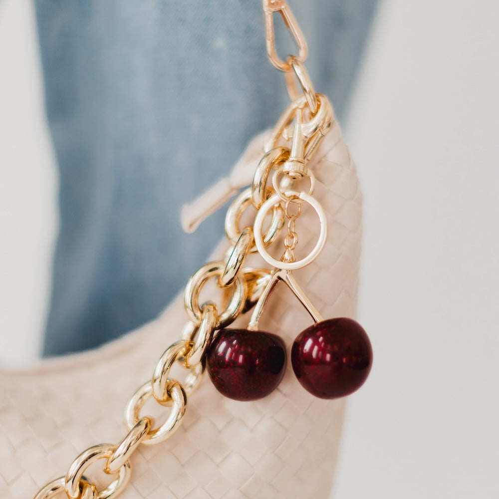 Gold chain with cherry-shaped charms on a beige handbag.