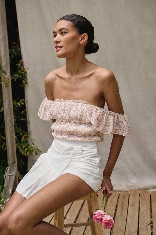 A female model wearing a floral square neck bodysuit with a gathered bodice and low back. The image shows the model seated, wearing white shorts and holding a flower.