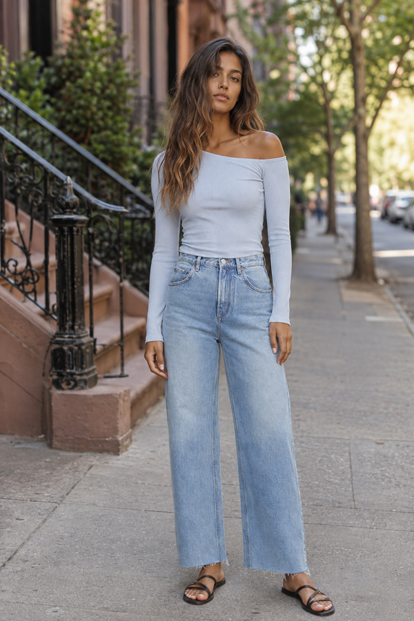 A woman stands on a city sidewalk wearing a light off-the-shoulder long-sleeve top and light-wash wide-leg jeans, paired with black sandals.