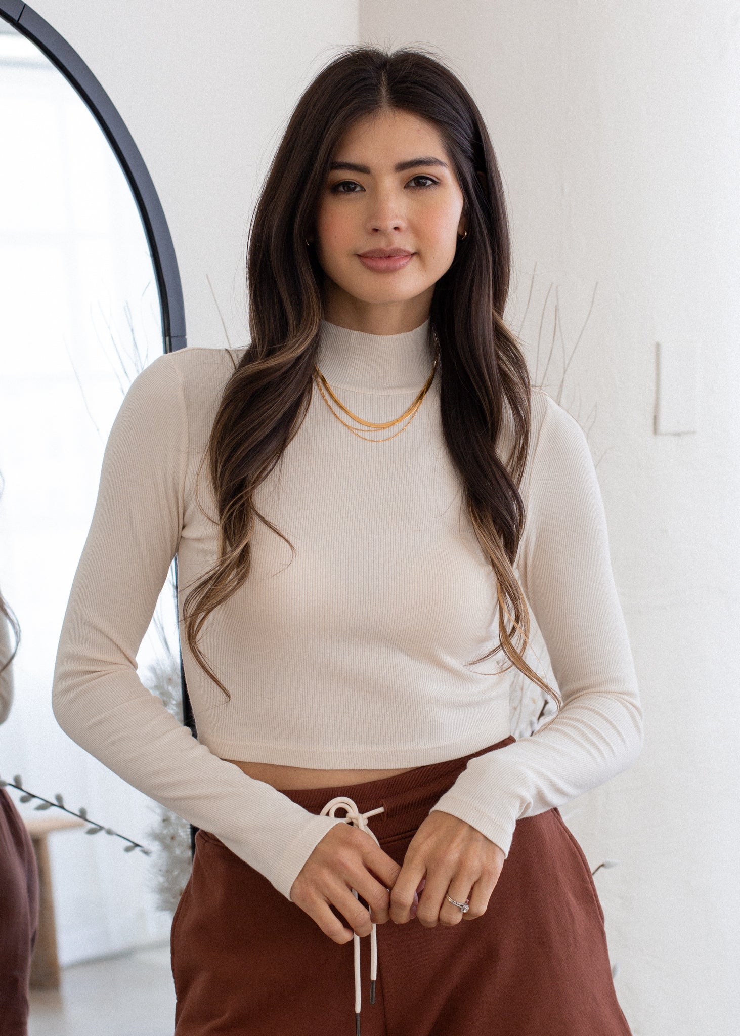 Woman wearing a black long-sleeve top and gray pants against a white wall.