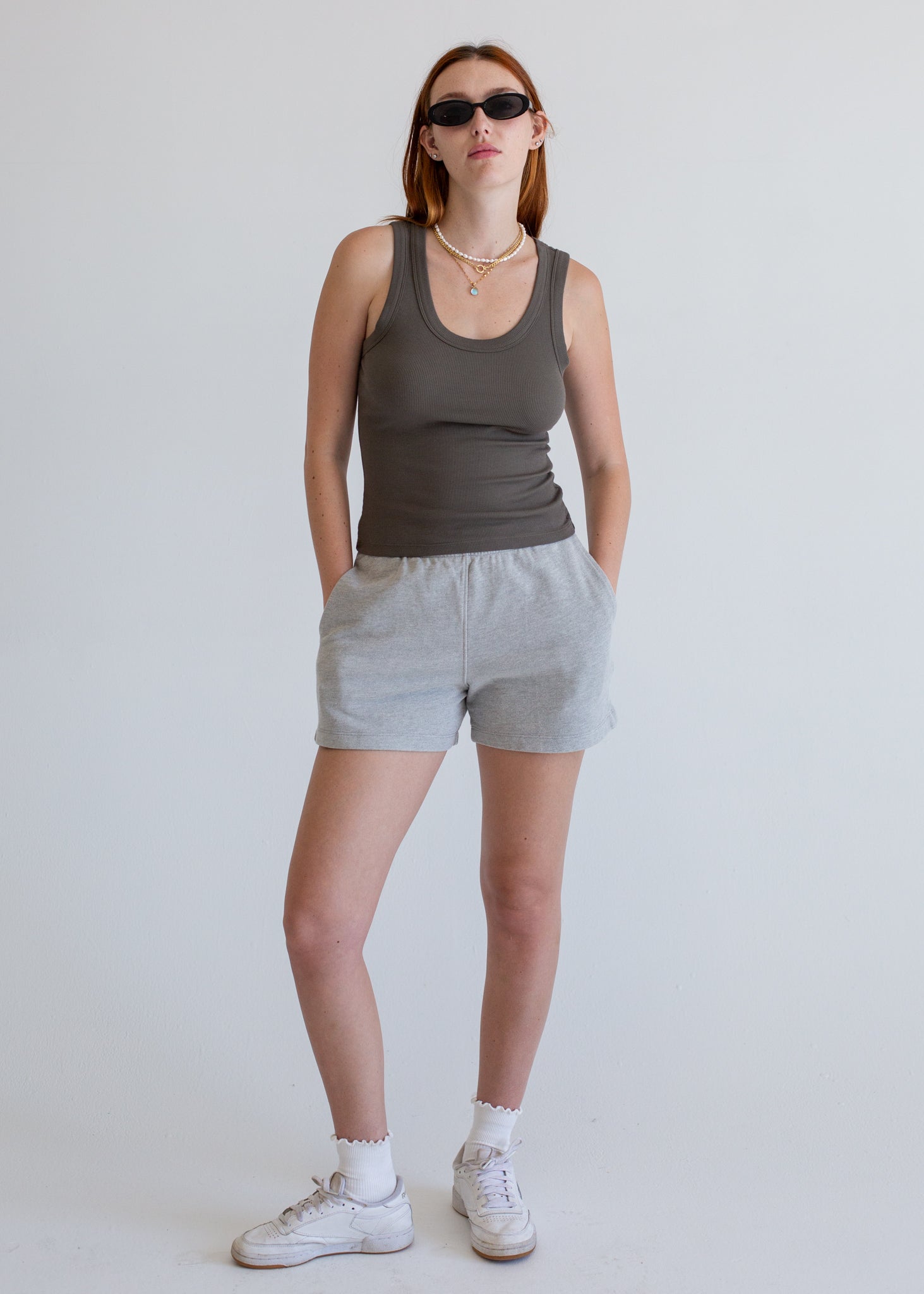 Woman wearing a white tank top and olive green shorts against a plain background