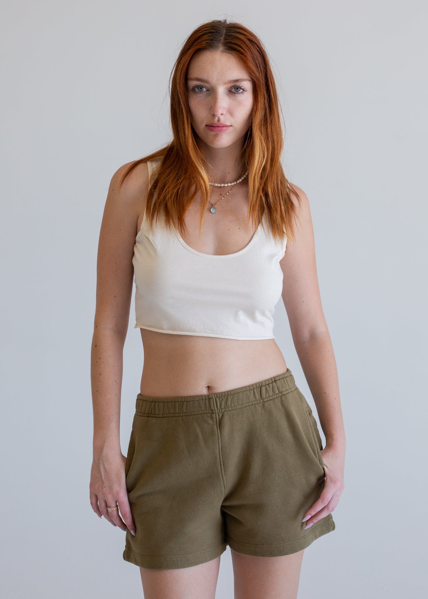 Woman wearing a white tank top and olive green shorts against a plain background