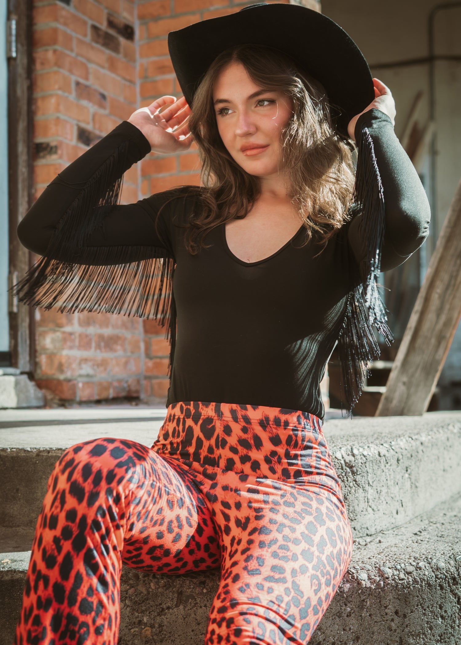 Sheer fringe bodysuit with dramatic fringe sleeves, plunging neckline, and cheeky cut – edgy night-out statement outfit for women, with a cowboy hat and orange and black animal print pants
