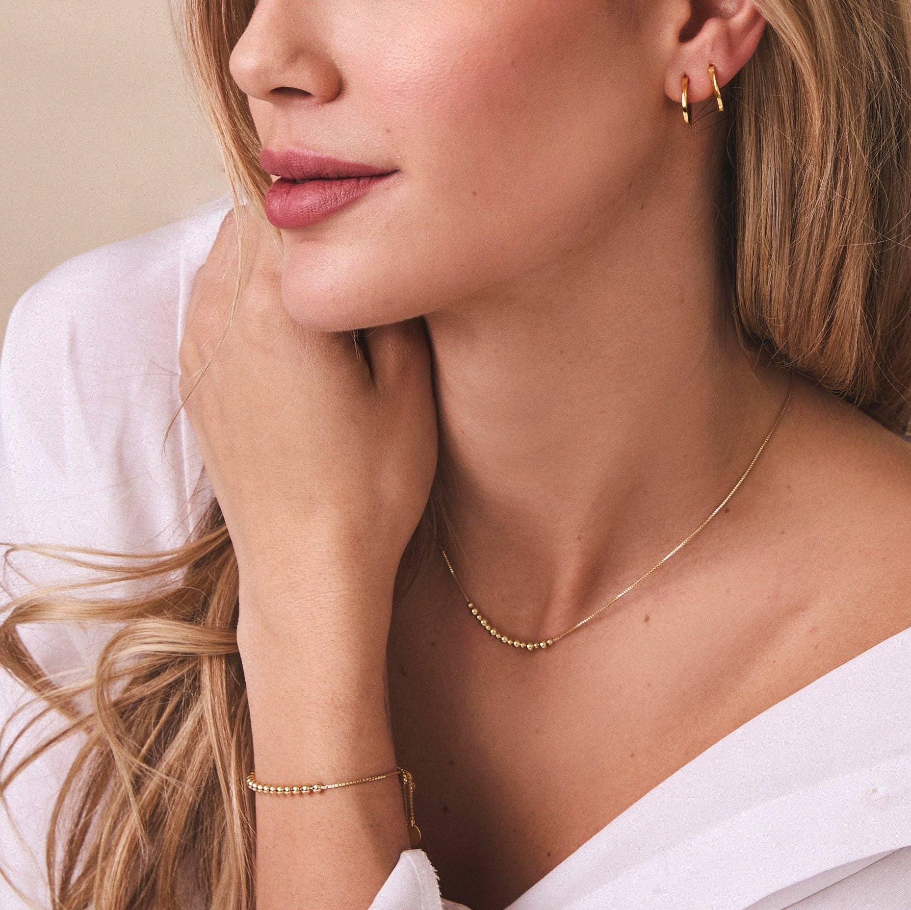 Close-up of a woman wearing gold jewelry including earrings, necklace, and bracelet on a neutral background.