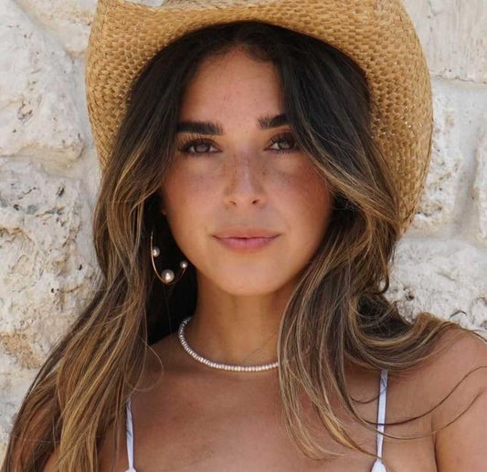 Woman wearing a straw hat and pearl necklace against a stone wall.