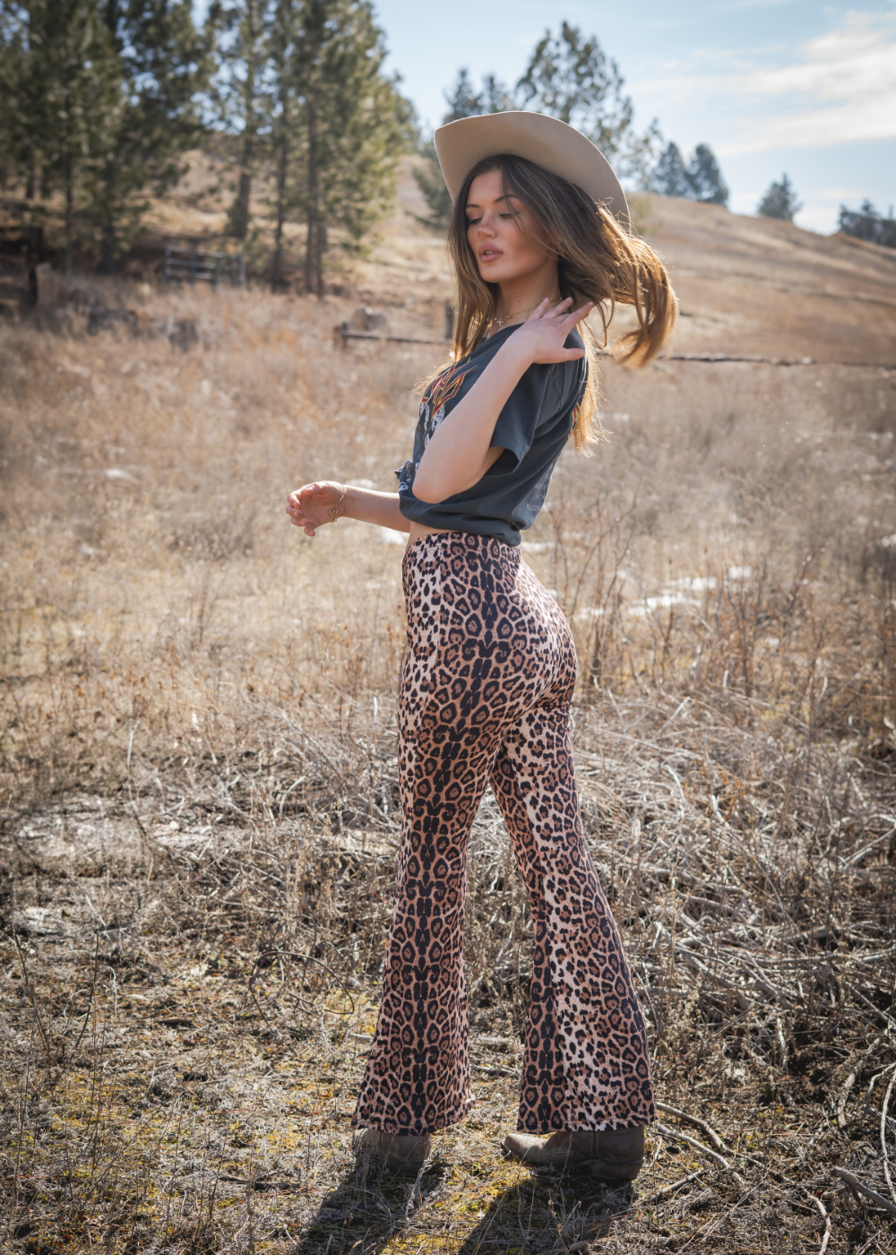 Woman in leopard print pants and cowboy hat standing in a field with trees in the background