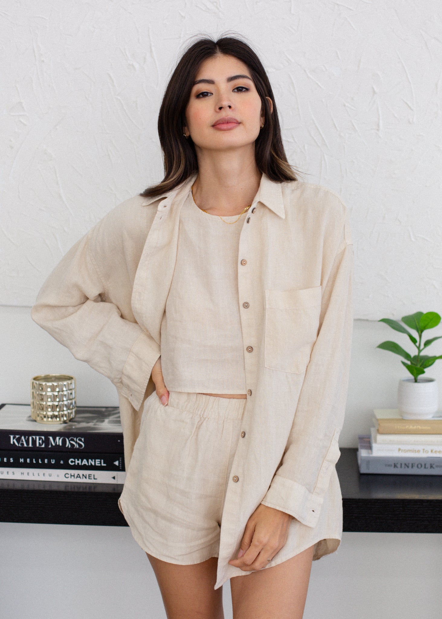 Woman wearing a beige outfit standing in front of a white wall with books and a plant.