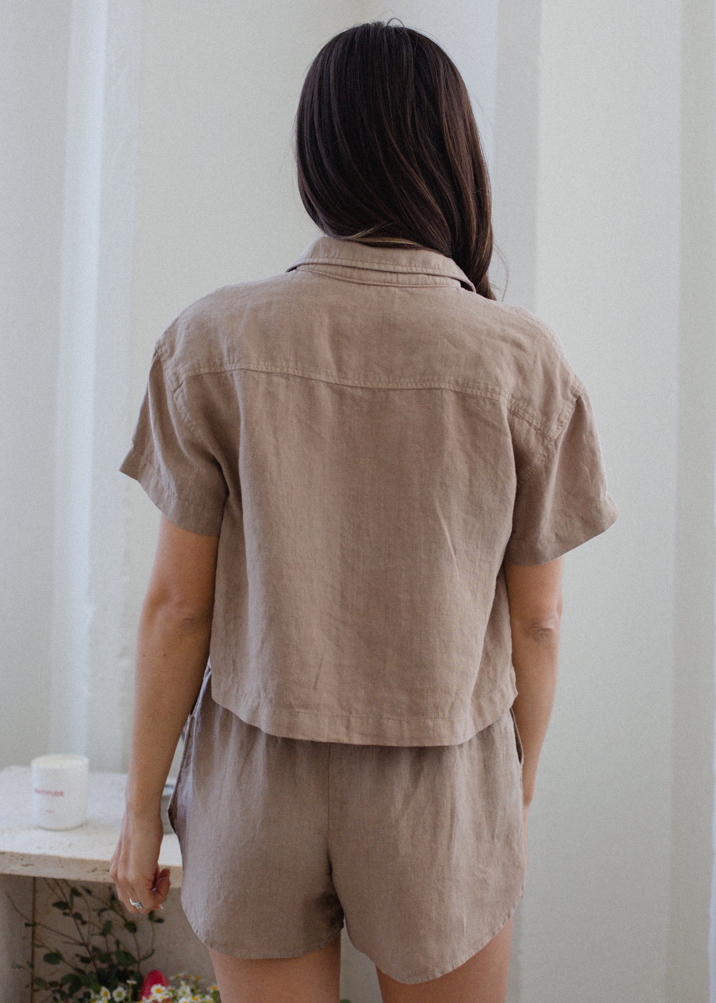 Person wearing a beige short-sleeve shirt and shorts set in a minimal indoor setting.